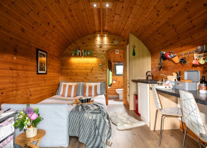 Inside a wooden glamping pod, looking over a cosy kingsize bed and a well-equipped kitchenette, with a view through an open door to a bathroom