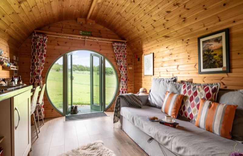 A view over a cosy sofa and well-equipped kitchenette out of a round door onto green fields beyond