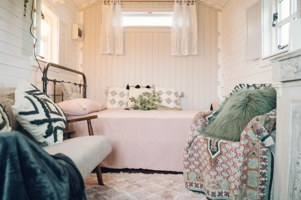 A cozy bedroom with white wooden walls, a metal bed frame with pink bedding, and decorative pillows. There is a small sofa with a patterned pillow and an animal print blanket, along with lush green plants on the bed. The room has natural light coming