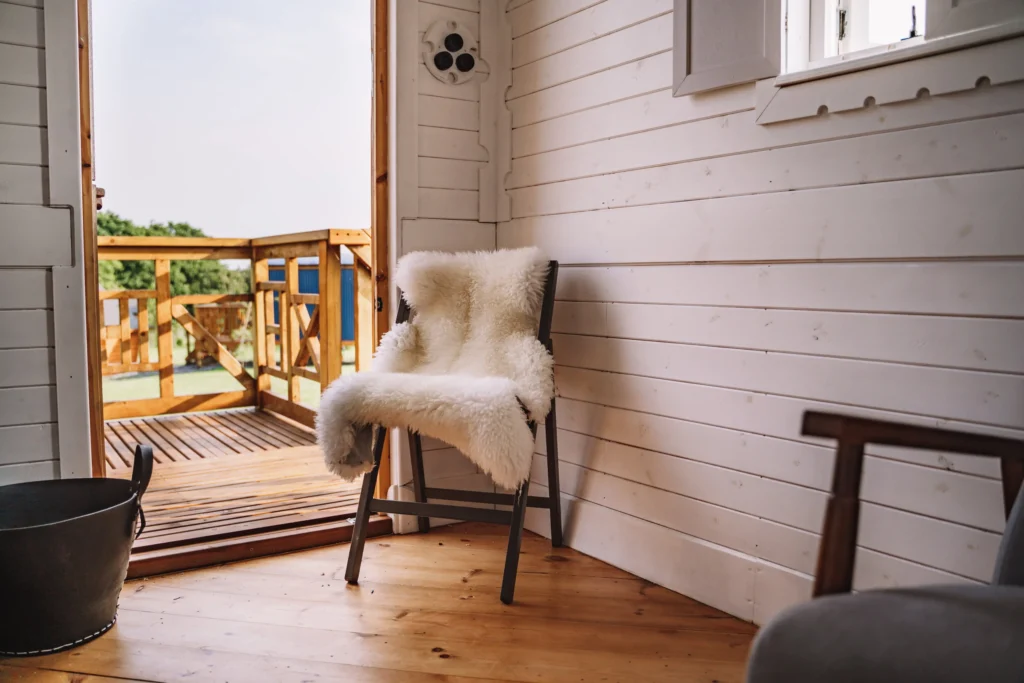 Interior of a cozy room with white paneled walls and a wooden floor, featuring a chair draped with a fluffy white throw, next to an open door leading to a wooden balcony outdoors.