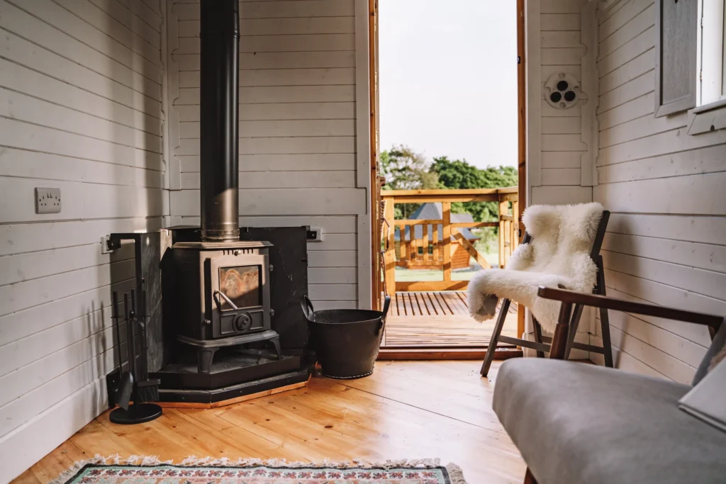 Cozy indoor space with a wood stove, a black bucket, a white fluffy blanket on a chair, and an open door leading to a wooden balcony with trees in the background.