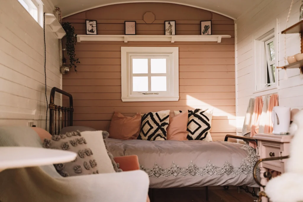 Cozy bedroom with a pink and white color scheme, featuring a bed with multiple pillows, a white side table with a kettle, and framed artwork above a window on wood-paneled walls. Shepherds Hut Glamping near Eryri / Snowdonia National Park.