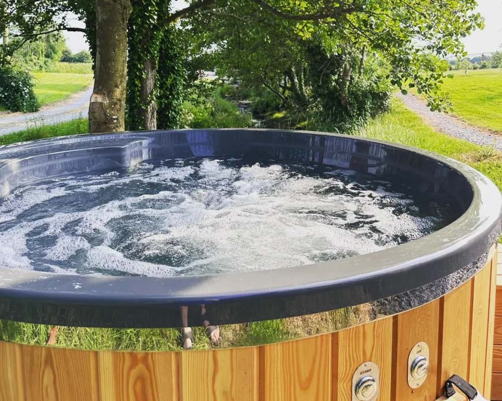 Hot tub with bubbling water outdoors on a grassy area with trees and a gravel path in the background. Hot Tub Glamping near Snowdonia National Park. Eryri.